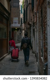 Venice, Italy - May 3 2019: A Unidentified Woman Tourist With Red Rolling Suitcase And Man With Backpack Walking Along The Narrow Street In Venice Back View