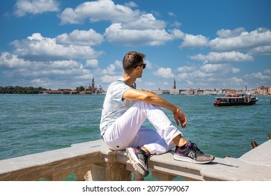 Venice, Italy. September 15, 2021. Handsome male tourist sitting on concrete fenced embankment in front of sea with cityscape in the background