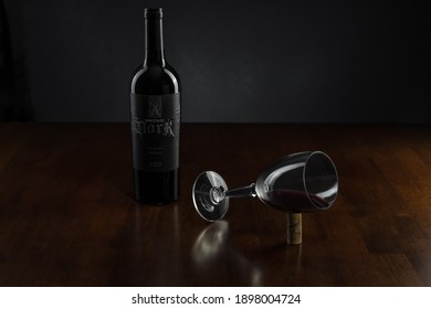 Vero Beach, Florida; USA; Dec. 7, 2020. A Bottle Of Apothic Dark Red Wine Blend Accompanies A Wine Glass Tilted Onto Its Side At The Center Of A Rosewood Table. The Image Has Dark And Moody Lighting. 