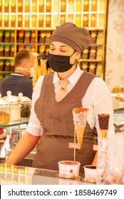 Verona, Veneto/Italy - 18.08.2020: An Ice Cream Saleswoman In An Italian Ice Cream Parlor In Brown Uniform And A Protective Mask In Front Of Her Various Kinds Of Ice Cream And The Waffles And Cups