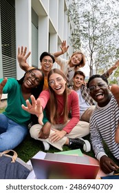 Vertical. Amusing portrait of group diverse young friends excited waving at camera with hands sitting on campus grass. Gen z of happy students gathered posing together for photo outside faculty class