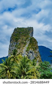 Vertical angle of an ancient karst limestone tower rising above tropical palm trees under a dramatic cloudy sky.
