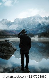 A vertical back view of man with fedora hat standing on rock near lake