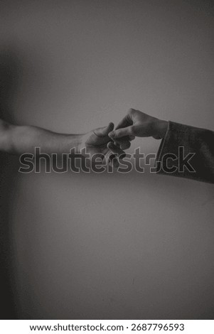 Vertical black and white close up of two hands gently touching fingers against a neutral wall. A conceptual image of human connection, intimacy, support, and fragile hope.