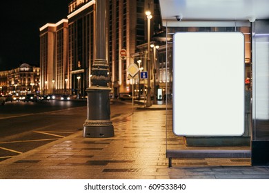 Vertical Blank Glowing Billboard At Bus Stop On The Night City Street. In The Background Buildings And Road With Cars. Mock Up. The Poster On The Street Next To The Roadway.