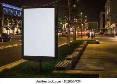 Vertical Blank Glowing Billboard On Night City Street. In Background Buildings And Road With Cars. Mock Up. Light Box On Street Next To Roadway. Space For Logo, Text, Image, Advertising, Ad, Blurb.