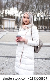 Vertical Blond Attractive Woman, Staying Look At Camera, In Warm White Jacket, Leather Bag Pack, Drinking Cup Of Coffee, Trees Park Plants Road. Urban Touristic Center, Weekend Activity, Joy