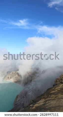 Vertical capture of steep volcanic cliffs bordering a turquoise crater lake under rising sulfur steam and a blue sky