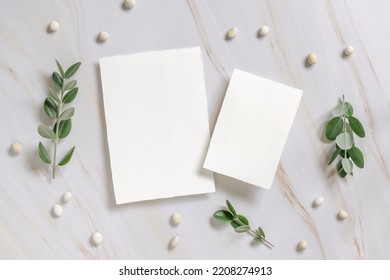 Vertical Cards On A Marble Table Decorated With Eucalyptus Branches And White Stones Top View, Wedding Mockup. Romantic Flat Lay With Blank Menu Card. Minimal Composition. Close To Nature 