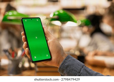 Vertical chroma key editable screen on smartphone held by person at library, green screen markers during study group, Mobile app mockup space with blurred green lamps.