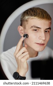 Vertical Close Up Portrait Of Blonde Young Man Using Face Massager While Filming Male Skincare Tutorial With Ring Light