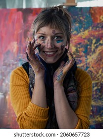 Vertical Close Up Portrait Of A Caucasian Female Artist Covered In Paint, Looking Directly At The Camera, Smiling. Standing In Front Of Her Modern Abstract Art Piece In An Empty Warehouse Space. 