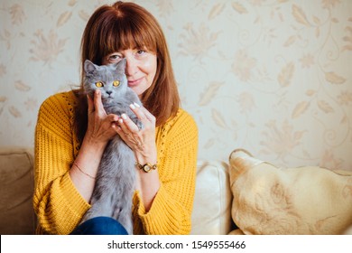 Vertical Close Up Portrait Of Lovely Middle-aged Redhead Woman With Gray Cat At Home. Mature Female In Cozy Yellow Sweater Cover Her Face With Cute Pet. Friendship Pet Lover Concept.