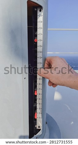 Vertical close up of a sailor hand adjusting a metallic measurement scale instrument with red indicators on the deck of a commercial ship sailing at sea.