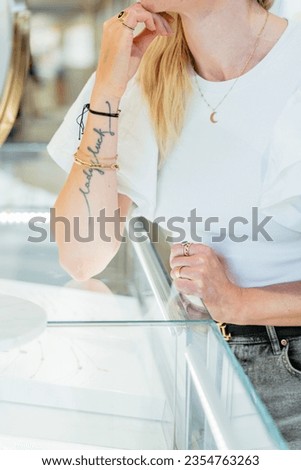Vertical close up shot of unrecognizable blond woman in white t-shirt looking at mirror try golden ring, bracelet, earrings in jewelry boutique.