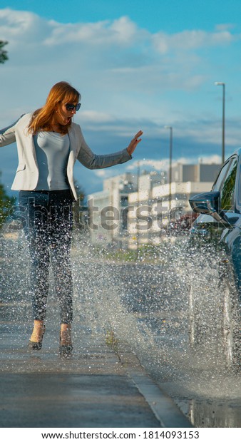 VERTICAL, CLOSE UP, DOF: Young businesswoman is\
surprised as she gets splashed with water by careless driver.\
Shocked female pedestrian gets splashed with dirty rainwater by SUV\
driving into a\
puddle.
