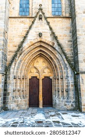 Vertical close view of a historic gothic church portal in Amberg, Upper Palatinate, Bavaria, Germany. The stone facade features an ornate pointed arch with carved details and a pair of tall dark wooden doors. Snow and wet cobblestones cover the ground in front of the entrance, captured in natural daylight.