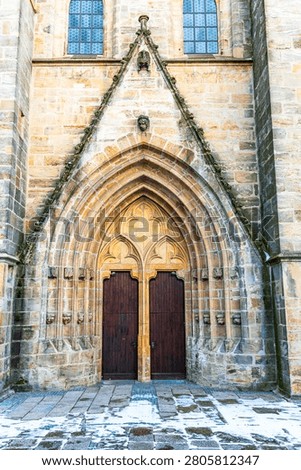 Vertical close view of a historic gothic church portal in Amberg, Upper Palatinate, Bavaria, Germany. The stone facade features an ornate pointed arch with carved details and a pair of tall dark wooden doors. Snow and wet cobblestones cover the ground in front of the entrance, captured in natural daylight.