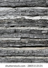 Vertical close up of weathered log cabin wall logs with flat split cracked wood, raised grain and several bent nails. The thick uneven chinking fills the gaps between the logs, with cracked and chipped areas exposing open spaces.