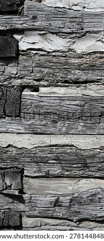 A vertical close up of weathered square logs and chinking from a log cabin, showing exposed natural patina wood with cracks, knots, splintering, and uneven surfaces. The porous cracked chinking fills the gaps between the logs, with rusty nail heads, nail holes, and small pebbles visible under even lighting.
