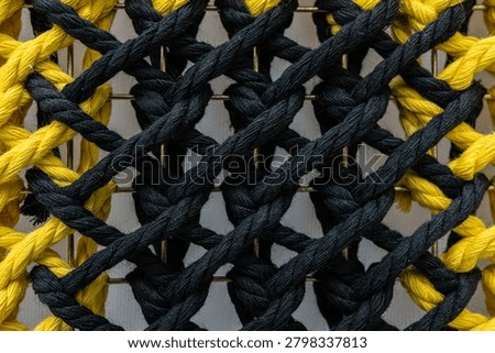 Vertical close-up of a decorative textile featuring black and yellow braided cords woven in a repetitive pattern over a metallic grid. Ideal for handicrafts and design.