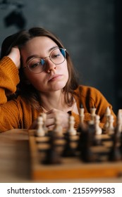 Vertical Close-up Face Of Pensive Young Woman In Elegant Eyeglasses Thinking About Chess Move Sitting In Dark Room, Selective Focus. Pretty Intelligent Lady Playing Logical Board Game Alone At Home.