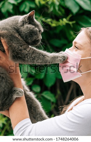 Vertical close-up portrait happy beautiful young woman with cat touching her pink face mask. Green leaves background. Concept of pet allergy or prevention of viral diseases