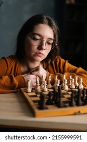 Vertical close-up portrait of pensive young woman in elegant eyeglasses thinking about chess move sitting in dark library room. Pretty intelligent lady playing logical board game alone at home. - Shutterstock ID 2155999591