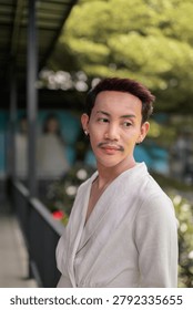 A vertical close-up portrait of a stylish non-binary gay Thai man with red-brown hair and a mustache, wearing a fashionable cream wrap cardigan dress jacket. He is standing outside under a covered pavilion or walkway structure, turning his head over his shoulder to look away thoughtfully to the side. The tight framing accentuates his facial expression, multiple ear piercings, and elegant linen-texture clothing. The background shows a beautifully soft, shallow depth of field blur featuring lush green trees, garden foliage, dark structural support pillars, and a metal railing. This authentic, unaltered image captures contemporary LGBTQ+, queer, and gender-fluid identity alongside themes of urban lifestyle, contemplation, and modern fashion in Southeast Asia.