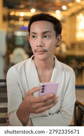 A vertical close-up portrait of a stylish non-binary gay Thai man with styled red-brown hair and a mustache, dressed in a fashionable cream wrap cardigan jacket. He is sitting outside a brightly lit commercial building or cafe at night, holding a pink smartphone in the foreground while looking away to the side with a thoughtful, curious expression. The background features a very warm, beautiful bokeh with out-of-focus hanging decorative ceiling lights, a glowing neon sign, and soft window displays. This authentic, unaltered image captures contemporary LGBTQ+, queer, and gender-fluid representation, pensive mobile technology habits, and modern urban evening lifestyle in Southeast Asia.