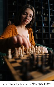 Vertical Close-up Portrait Of Young Woman In Elegant Eyeglasses Making Chess Move Sitting On Armchair In Dark Room, Selective Focus. Pretty Intelligent Lady Playing Logical Board Game Alone At Home.