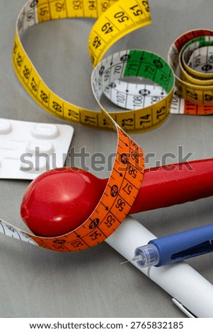 A vertical close-up shot featuring a red dumbbell with an orange measuring tape coiled around it, positioned alongside a blue insulin pen and a white blister pack of pills on a grey surface