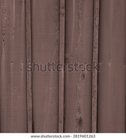 Vertical close-up view of a brown painted board and batten wood siding surface. The boards and batten are dried and have some cracks. The wood boards are stained where the nail head fasteners have rusted and run down the surface. The boards and batten have a rough, uneven texture with visible wood grain and knots. There is a splintered piece of misaligned unpainted batten near the bottom frame.