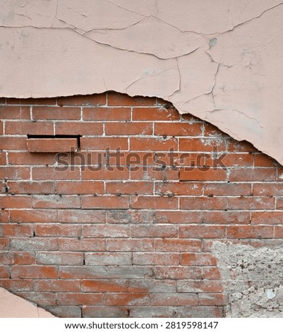 A vertical close-up view of a decaying multi-layer wall surface with tan painted concrete and an inner red brick layer with mortar. The tan painted concrete is scratched, cracked, and pulling away from the inner layers. The red brick layer is weatherworn with a misplaced brick sticking out, cracked and uneven mortar wearing away in places, and a large wall patch is visible in the lower corner.