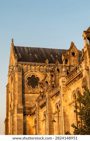 Vertical close-up view of the transept and upper nave of the neo-Gothic Sacré-Coeur church in Bordeaux, France, its soaring height emphasized against a clear blue sky in warm evening light. The image highlights intricate stonework, buttresses, and vertical architectural lines. No people present.