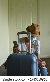 Vertical closeup of young Latin woman wearing jeans, white t-shirt and straw hat, with mask checking blue suitcase, leaving home, with copy space