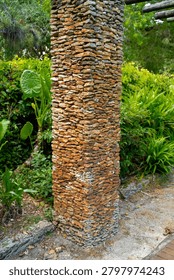 The vertical column consists of flat, brown, and yellow stacked stones near the green leaves of the tropical Alocasia plant in the botanical garden. The rough masonry construction block decorates the outdoor walking path for commercial architectural landscaping design and building projects.