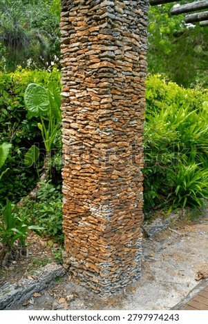 The vertical column consists of flat, brown, and yellow stacked stones near the green leaves of the tropical Alocasia plant in the botanical garden. The rough masonry construction block decorates the outdoor walking path for commercial architectural landscaping design and building projects.