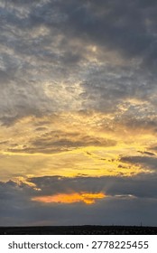 Vertical composition of dramatic sunset sky with glowing sunlight beneath dark storm clouds. Atmospheric evening landscape featuring layered cloud formations and warm golden horizon colors.