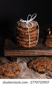 Vertical composition featuring stack of chocolate chip cookies tied with white string, placed on dark wooden riser. More cookies are scattered on the wooden table surface. Dusted with icing sugar against black background.