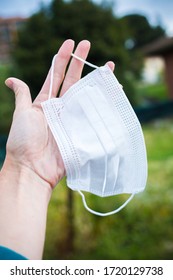 Vertical Composition. Medical Protective Mask In Woman’s Hand With Green Natural Background. COVID 19 Coronavirus Lifestyle Scene. No People Medical Outdoor Composition. Fingers Hold White Thing