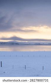 A vertical composition of a serene winter scene during sunset. A rustic wire fence stands in a deep snow field, with a frozen lake and snow-covered hills in the distance under a colorful golden sky.