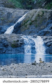 A vertical composition showcasing a multi-level waterfall cascading down dark cliffs into a pristine blue river basin with a pebble shore in the front