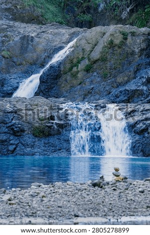 A vertical composition showcasing a multi-level waterfall cascading down dark cliffs into a pristine blue river basin with a pebble shore in the front