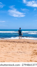 Vertical composition of a surfer standing on the shoreline seen from behind while looking at ocean waves in a calm coastal setting