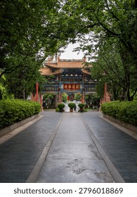 A vertical composition where dense green foliage frames a central walkway leading to a colorful traditional gate in the distance. The image represents sanctuary, hidden heritage, urban nature, and peaceful atmosphere.