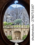 Vertical, creative shot looking through a circular stone window opening. The oval framing captures a centralized view of the elaborate Baroque-style stone structure featuring grand arched niches, classical statues, a central fountain, and lush green hedges. Tall, bare deciduous winter trees and rolling hills in the background complete the scenic landscape vista.