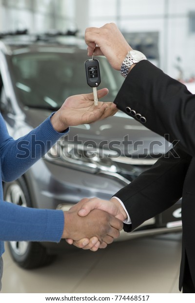 Vertical cropped close up of a car dealer and his
client shaking hands. Professional salesman giving car keys to his
customer sharing handshake cars on the background buying buyer
purchasing renting