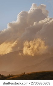 Vertical dramatic landscape with large fire clouds and small cabin building on grassy hill illuminated by warm golden light of sunrise or sunset.