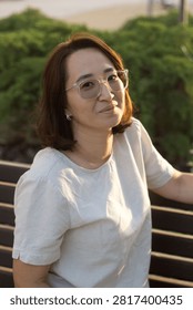 A vertical, eye-level medium portrait of a smiling mid-adult woman of Central Asian ethnicity. She is sitting comfortably on a dark slatted wooden park bench, leaning slightly forward with a gentle, confident expression while looking directly at the camera. Her hands rest naturally on her lap and knee. She has shoulder-length dark brown hair and wears small silver hoop earrings, a light cream-colored short-sleeve linen top, and dark grey trousers. The scene is illuminated by the soft, warm golden light of a late afternoon or sunset, casting bright highlights on her face and top. In the blurred background, a paved park path and low evergreen bushes sit under warm ambient light.